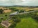 A steam train travels across a scenic countryside landscape, trailing white smoke behind it. The train tracks run parallel to lush green fields and trees. In the foreground, a cottage with a stone roof is visible, surrounded by gardens and a few scattered sheep.