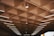 A ceiling with a geometric pattern of square recessed panels, each housing a small central light fixture. Linear fluorescent lights are integrated into the structure, providing parallel illumination across the surface. The lighting creates a warm atmosphere, with shadows accentuating the grid pattern.