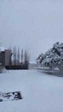 A snowy landscape featuring a yard covered in a thick layer of white snow. There is a fence running along one side, with trees in the background also blanketed in snow. A building with tall, thin trees at its side is visible, contributing to the wintry scene.