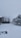A snowy landscape featuring a yard covered in a thick layer of white snow. There is a fence running along one side, with trees in the background also blanketed in snow. A building with tall, thin trees at its side is visible, contributing to the wintry scene.
