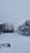 A snowy landscape featuring a yard covered in a thick layer of white snow. There is a fence running along one side, with trees in the background also blanketed in snow. A building with tall, thin trees at its side is visible, contributing to the wintry scene.