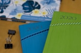 Two 2020 planners or notebooks are prominently displayed, one in blue and the other in green, each with a decorative striped pattern. A floral design is visible in the background on another item. A black binder clip is also seen on the wooden surface.