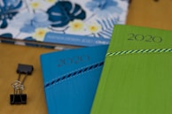 Two 2020 planners or notebooks are prominently displayed, one in blue and the other in green, each with a decorative striped pattern. A floral design is visible in the background on another item. A black binder clip is also seen on the wooden surface.