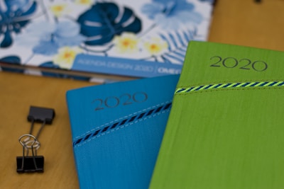 Two 2020 planners or notebooks are prominently displayed, one in blue and the other in green, each with a decorative striped pattern. A floral design is visible in the background on another item. A black binder clip is also seen on the wooden surface.