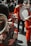 A group of people dressed in red military uniforms play musical instruments such as drums and brass horns. Their uniforms include tall, furry black hats, and the scene appears to be part of a ceremonial or parade event.