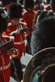 A group of uniformed musicians stand in formation, wearing red coats with gold embellishments and tall black fur hats. They hold brass instruments and appear to be part of a formal marching band.