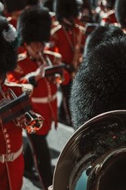 A group of uniformed musicians stand in formation, wearing red coats with gold embellishments and tall black fur hats. They hold brass instruments and appear to be part of a formal marching band.