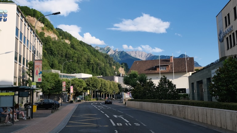 A street view in a small town is depicted with a clear road leading towards a distant mountain range. The area is flanked by modern buildings, including an office labeled with 'VP Bank.' Green trees line the street, and a bus stop with people sitting is visible on the left side. The sky above is blue with scattered clouds.