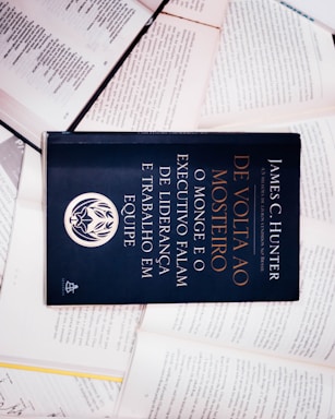 A dark-colored book titled 'De Volta ao Mosteiro' by James C. Hunter is lying on top of several open books with pages of text visible. The book cover features gold and white text along with an emblem or logo in white.