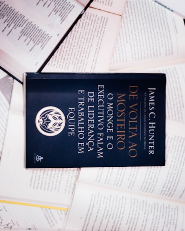 A dark-colored book titled 'De Volta ao Mosteiro' by James C. Hunter is lying on top of several open books with pages of text visible. The book cover features gold and white text along with an emblem or logo in white.
