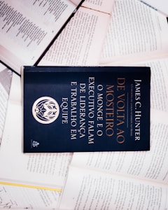 A dark-colored book titled 'De Volta ao Mosteiro' by James C. Hunter is lying on top of several open books with pages of text visible. The book cover features gold and white text along with an emblem or logo in white.
