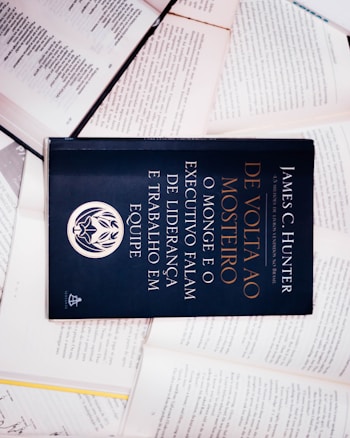 A dark-colored book titled 'De Volta ao Mosteiro' by James C. Hunter is lying on top of several open books with pages of text visible. The book cover features gold and white text along with an emblem or logo in white.