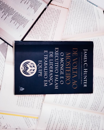 A dark-colored book titled 'De Volta ao Mosteiro' by James C. Hunter is lying on top of several open books with pages of text visible. The book cover features gold and white text along with an emblem or logo in white.