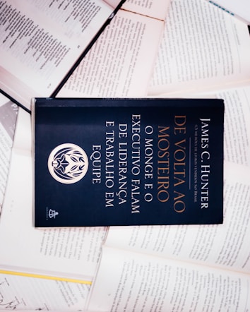 A dark-colored book titled 'De Volta ao Mosteiro' by James C. Hunter is lying on top of several open books with pages of text visible. The book cover features gold and white text along with an emblem or logo in white.