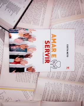 A book titled 'Amar e Servir' with an image of diverse hands reaching upwards is placed on top of several open books. The scene appears to be well-lit, highlighting the book cover prominently.