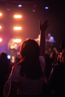 A scene of a concert or live event with a person in the foreground, silhouetted against bright stage lights. The hand is raised, conveying enthusiasm and engagement with the performance. Other audience members are visible facing the stage where performers are slightly blurred.