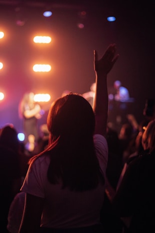A scene of a concert or live event with a person in the foreground, silhouetted against bright stage lights. The hand is raised, conveying enthusiasm and engagement with the performance. Other audience members are visible facing the stage where performers are slightly blurred.