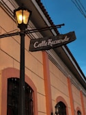 A warm, illuminated street sign hangs from an old-fashioned street lamp on a narrow street. The sign reads 'Calle Recuerdo' and is attached to a building with beige and orange tones. The architectural style suggests a historic setting, and the sunset light casts a soft blue hue in the sky.