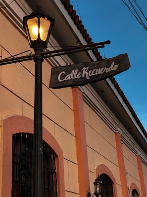A warm, illuminated street sign hangs from an old-fashioned street lamp on a narrow street. The sign reads 'Calle Recuerdo' and is attached to a building with beige and orange tones. The architectural style suggests a historic setting, and the sunset light casts a soft blue hue in the sky.