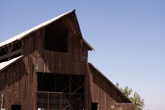 A large wooden barn structure with an A-frame roof, built using weathered wooden planks. The roof is partially covered with metal sheets, and sunlight creates shadows on the building. In the background, a clear blue sky stretches across the horizon with a few leafy green trees visible near the bottom right of the image.