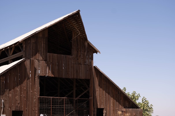 A large wooden barn structure with an A-frame roof, built using weathered wooden planks. The roof is partially covered with metal sheets, and sunlight creates shadows on the building. In the background, a clear blue sky stretches across the horizon with a few leafy green trees visible near the bottom right of the image.