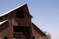 A large wooden barn structure with an A-frame roof, built using weathered wooden planks. The roof is partially covered with metal sheets, and sunlight creates shadows on the building. In the background, a clear blue sky stretches across the horizon with a few leafy green trees visible near the bottom right of the image.