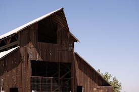 A large wooden barn structure with an A-frame roof, built using weathered wooden planks. The roof is partially covered with metal sheets, and sunlight creates shadows on the building. In the background, a clear blue sky stretches across the horizon with a few leafy green trees visible near the bottom right of the image.