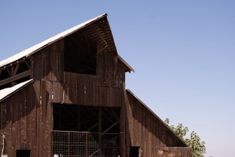 A large wooden barn structure with an A-frame roof, built using weathered wooden planks. The roof is partially covered with metal sheets, and sunlight creates shadows on the building. In the background, a clear blue sky stretches across the horizon with a few leafy green trees visible near the bottom right of the image.