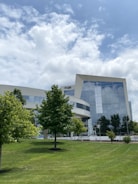 A modern office building with a sleek glass facade is surrounded by a green, manicured lawn and trees under a partly cloudy sky. The building features reflective windows, and its geometric design adds to the contemporary appearance. There are a few scattered trees in front of the structure and a small number of pedestrians in the distance.