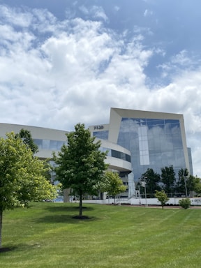 A modern office building with a sleek glass facade is surrounded by a green, manicured lawn and trees under a partly cloudy sky. The building features reflective windows, and its geometric design adds to the contemporary appearance. There are a few scattered trees in front of the structure and a small number of pedestrians in the distance.