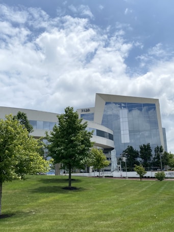 A modern office building with a sleek glass facade is surrounded by a green, manicured lawn and trees under a partly cloudy sky. The building features reflective windows, and its geometric design adds to the contemporary appearance. There are a few scattered trees in front of the structure and a small number of pedestrians in the distance.