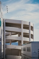 A multi-story parking structure with curved ramps is visible, set against a backdrop of clear blue sky. The architecture is modern and minimalist with white concrete surfaces. In the foreground, part of a building with a bicycle symbol can be seen, suggesting parking or city infrastructure.