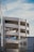 A multi-story parking structure with curved ramps is visible, set against a backdrop of clear blue sky. The architecture is modern and minimalist with white concrete surfaces. In the foreground, part of a building with a bicycle symbol can be seen, suggesting parking or city infrastructure.