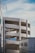 A multi-story parking structure with curved ramps is visible, set against a backdrop of clear blue sky. The architecture is modern and minimalist with white concrete surfaces. In the foreground, part of a building with a bicycle symbol can be seen, suggesting parking or city infrastructure.