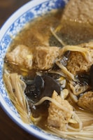 A close-up view of a bowl containing a broth with various ingredients including tofu, black fungus, and enoki mushrooms. The dish appears to have a rich and savory look with a clear, flavorful broth. The bowl is decorated with a blue-and-white pattern.