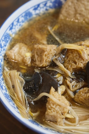 A close-up view of a bowl containing a broth with various ingredients including tofu, black fungus, and enoki mushrooms. The dish appears to have a rich and savory look with a clear, flavorful broth. The bowl is decorated with a blue-and-white pattern.