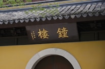 A traditional structure with Chinese characters in gold displayed on a sign above a rounded doorway. The building features a classic tiled roof with a slight curvature and is adorned with greenery.