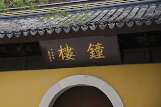 A traditional structure with Chinese characters in gold displayed on a sign above a rounded doorway. The building features a classic tiled roof with a slight curvature and is adorned with greenery.
