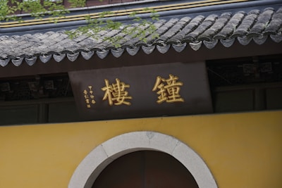 A traditional structure with Chinese characters in gold displayed on a sign above a rounded doorway. The building features a classic tiled roof with a slight curvature and is adorned with greenery.