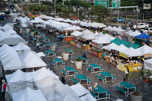 An open-air market with multiple white tents arranged in rows, offering a variety of goods and food items. Many of the stalls have colorful signage. In front of the tents are teal picnic tables with attached benches. Few people are visible walking or standing around the market area. There are also large potted plants spaced between the tables. The surrounding area includes a busy street with visible cars and lush green trees in the background.