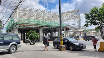 A large construction site features a structure with a metal framework and scaffolding. Vehicles are parked on the street, including a black car and a blue SUV. A person holding an umbrella walks along the sidewalk, while another person stands nearby carrying a backpack. Overhead, numerous power lines crisscross the sky, which is partly cloudy.
