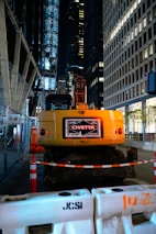 A construction site at night located in an urban area, featuring a yellow excavator with the label 'John Civetta' on its rear. The scene is surrounded by tall, illuminated office buildings. The area is cordoned off with construction barriers and traffic cones, highlighting the ongoing work. Metal fencing lines one side, adding to the industrial atmosphere.
