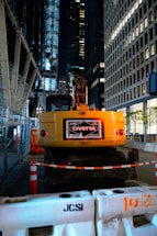 A construction site at night located in an urban area, featuring a yellow excavator with the label 'John Civetta' on its rear. The scene is surrounded by tall, illuminated office buildings. The area is cordoned off with construction barriers and traffic cones, highlighting the ongoing work. Metal fencing lines one side, adding to the industrial atmosphere.