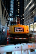 A construction site at night located in an urban area, featuring a yellow excavator with the label 'John Civetta' on its rear. The scene is surrounded by tall, illuminated office buildings. The area is cordoned off with construction barriers and traffic cones, highlighting the ongoing work. Metal fencing lines one side, adding to the industrial atmosphere.