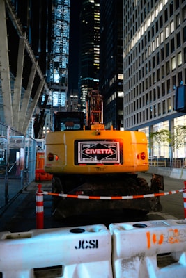 A construction site at night located in an urban area, featuring a yellow excavator with the label 'John Civetta' on its rear. The scene is surrounded by tall, illuminated office buildings. The area is cordoned off with construction barriers and traffic cones, highlighting the ongoing work. Metal fencing lines one side, adding to the industrial atmosphere.