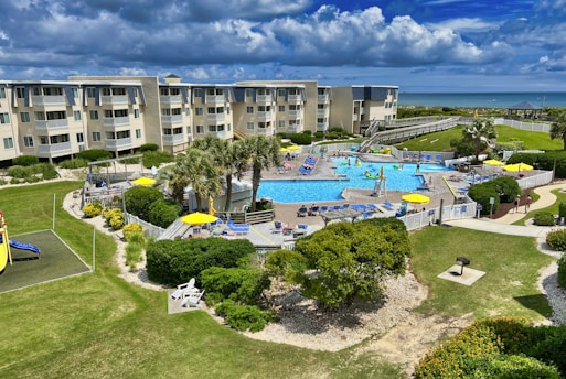 A coastal resort scene featuring a large swimming pool surrounded by sun loungers and umbrellas. Adjacent to the pool is a multi-story building with balconies overlooking the area. The landscape includes well-maintained lawns, various plants, and a playground. In the background, the ocean and a sandy beach are visible under a partly cloudy sky.