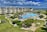 A coastal resort scene featuring a large swimming pool surrounded by sun loungers and umbrellas. Adjacent to the pool is a multi-story building with balconies overlooking the area. The landscape includes well-maintained lawns, various plants, and a playground. In the background, the ocean and a sandy beach are visible under a partly cloudy sky.