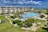 A coastal resort scene featuring a large swimming pool surrounded by sun loungers and umbrellas. Adjacent to the pool is a multi-story building with balconies overlooking the area. The landscape includes well-maintained lawns, various plants, and a playground. In the background, the ocean and a sandy beach are visible under a partly cloudy sky.