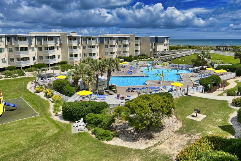 A coastal resort scene featuring a large swimming pool surrounded by sun loungers and umbrellas. Adjacent to the pool is a multi-story building with balconies overlooking the area. The landscape includes well-maintained lawns, various plants, and a playground. In the background, the ocean and a sandy beach are visible under a partly cloudy sky.