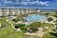 A coastal resort scene featuring a large swimming pool surrounded by sun loungers and umbrellas. Adjacent to the pool is a multi-story building with balconies overlooking the area. The landscape includes well-maintained lawns, various plants, and a playground. In the background, the ocean and a sandy beach are visible under a partly cloudy sky.
